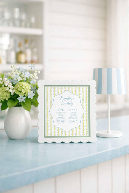a white framed sign with a green and white striped border, placed on a blue countertop next to a vase of white flowers and a white lamp.