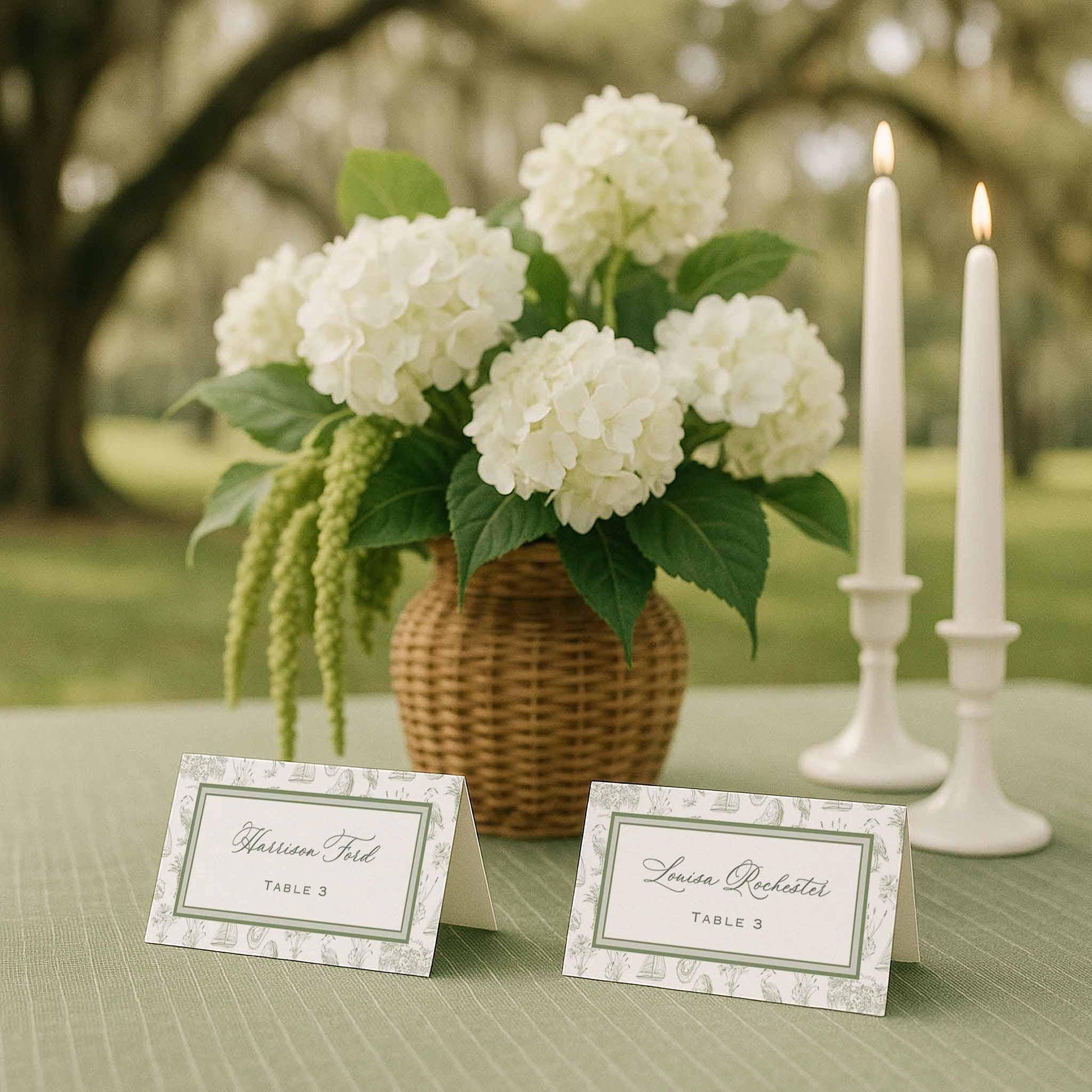Two folded Southern toile escort cards displayed on a sage green textured tablecloth beside a rattan vase filled with white and green hydrangeas. The cards feature a fine art toile border with hand-drawn coastal illustrations including palmettos, egrets, oysters, marsh grasses, oak trees, and sailboats. White taper candles stand in the background beneath oak trees draped in Spanish moss. Elegant outdoor southern wedding seating card arrangement.