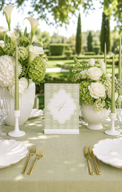 a beautifully set outdoor table for a wedding reception, with white flowers, candles, and place settings arranged on a green tablecloth.