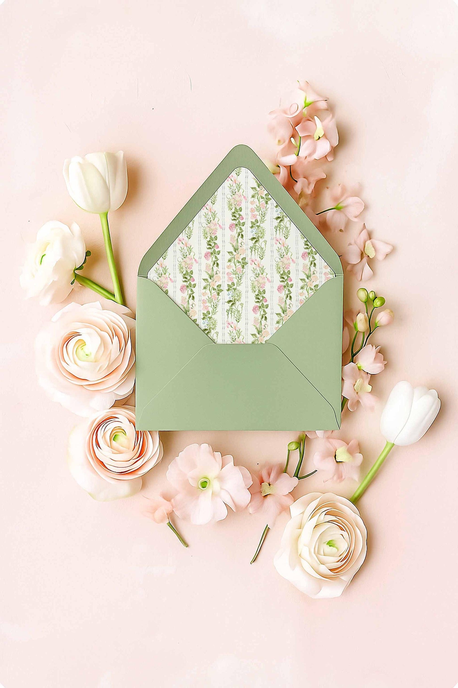 Green envelope with a floral watercolor envelope liner featuring hand-painted pink and green flowers, displayed in a flat lay with soft blush ranunculus, tulips, and florals on a pastel pink background.