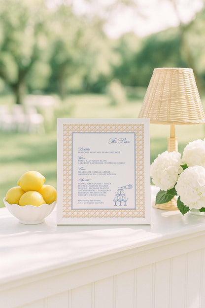a white table with a framed menu, a bowl of lemons, a lamp, and some flowers.