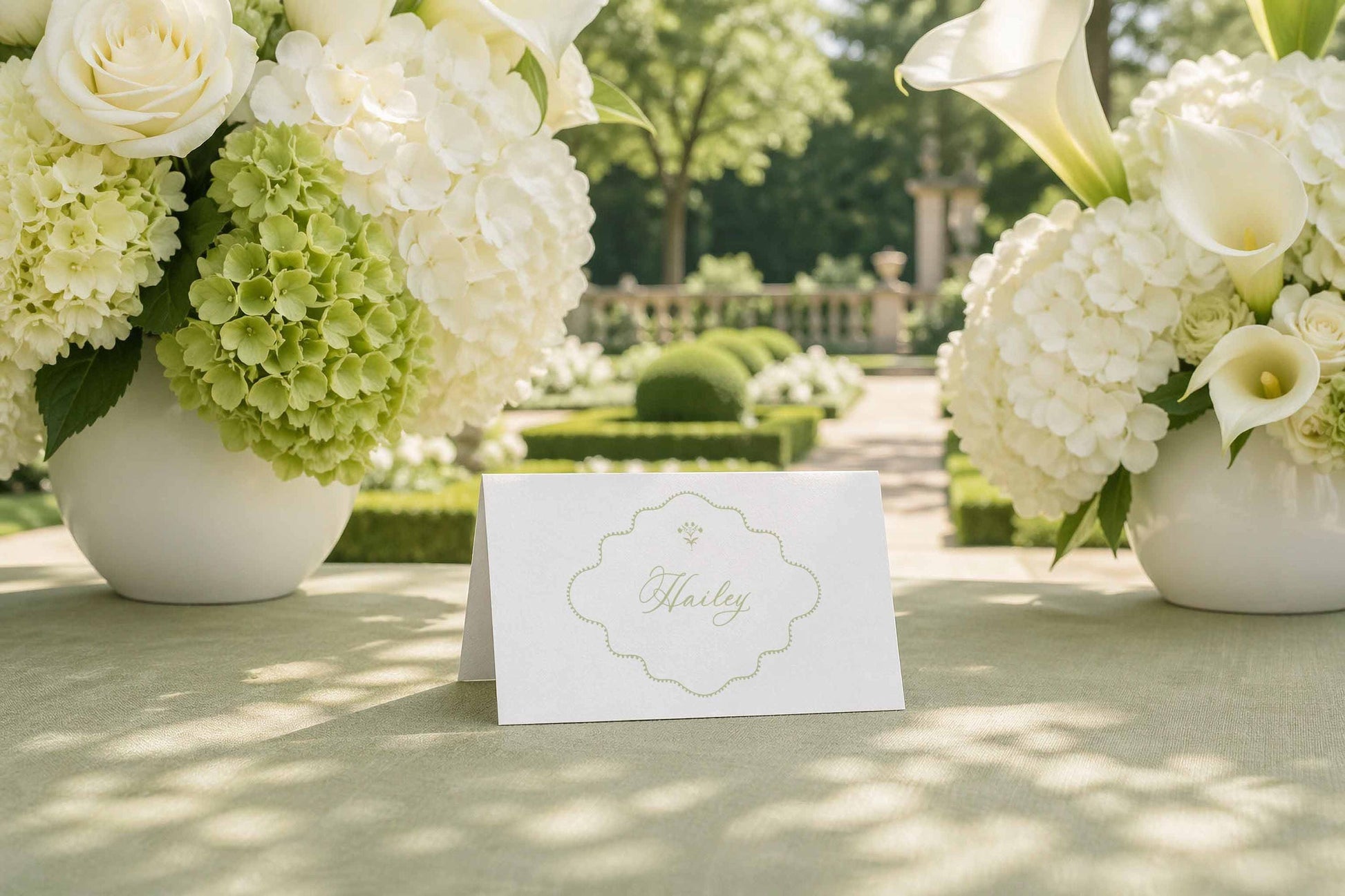 a table with a white card in the center, surrounded by two large vases filled with white flowers, likely roses, and a lush garden in the background.