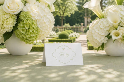 a table with a white card in the center, surrounded by two large vases filled with white flowers, likely roses, and a lush garden in the background.