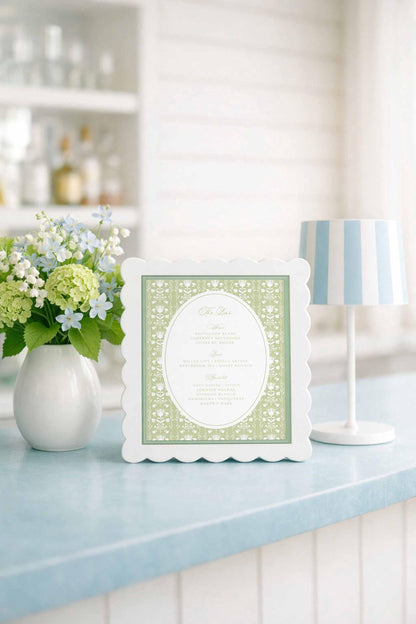 a white frame with a green floral design, placed on a blue countertop next to a vase of white and green flowers.