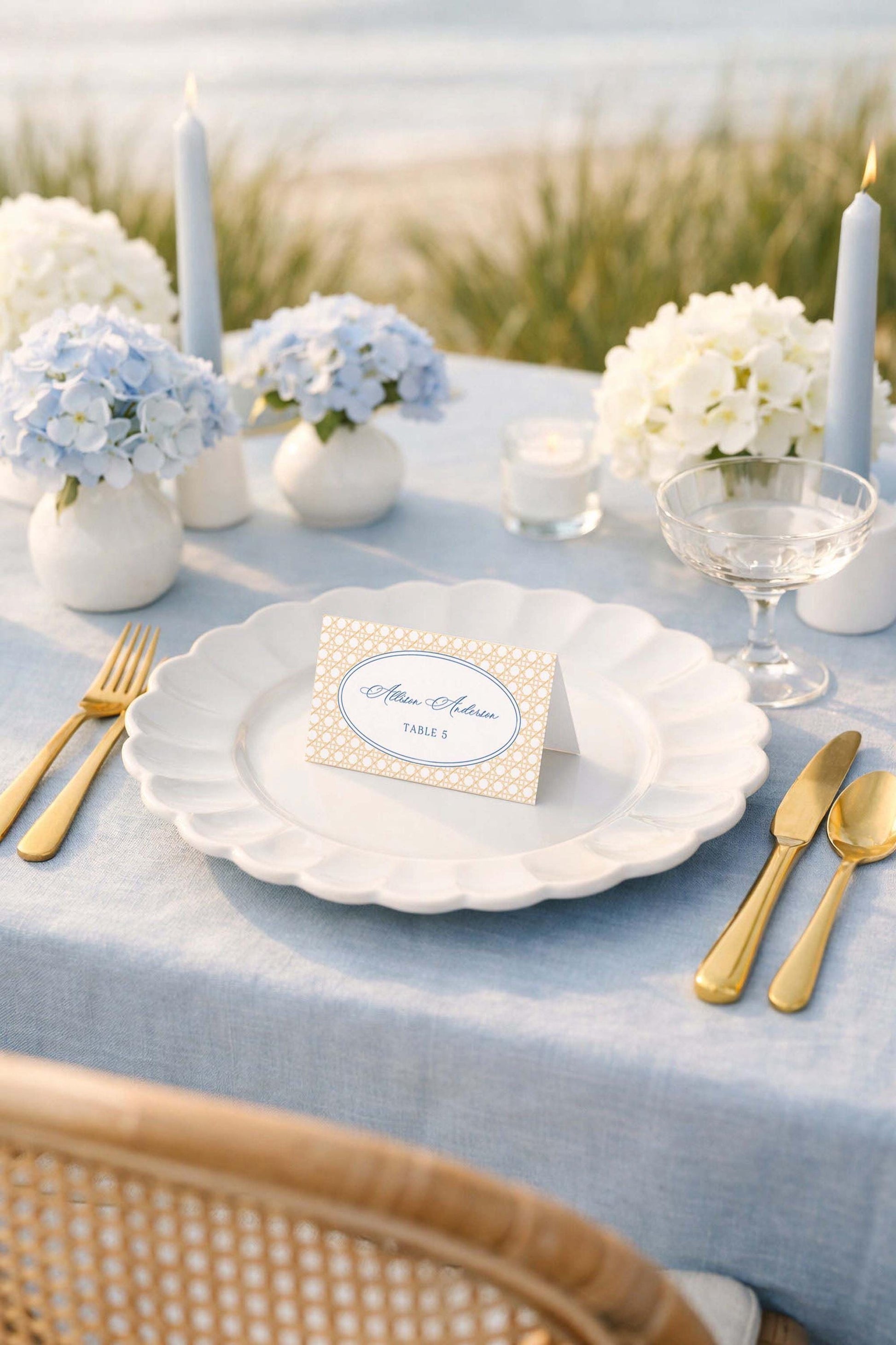 a beautifully set table for a wedding reception, with a blue tablecloth, white plates, gold cutlery, and a small card with the name 'Michele & Robert' on it. The table is set against a scenic backdrop of a beach, creating a romantic and elegant atmosphere.