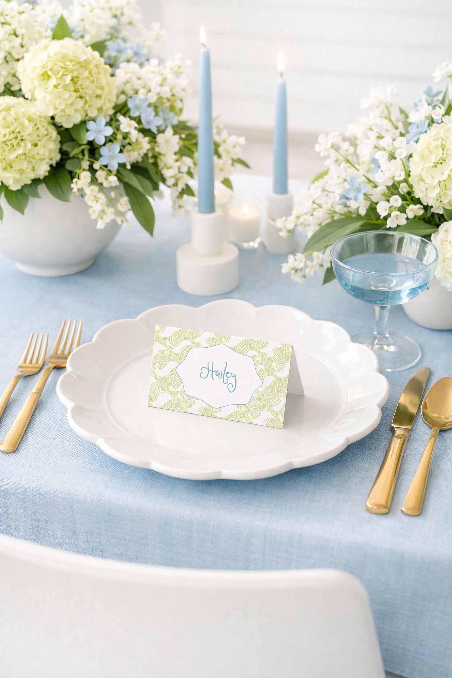 a beautifully set dining table with a white plate, gold cutlery, and a green and white place card with the name "Harley" on it. The table is adorned with white flowers, blue candles, and a glass of water, creating an elegant and inviting atmosphere.