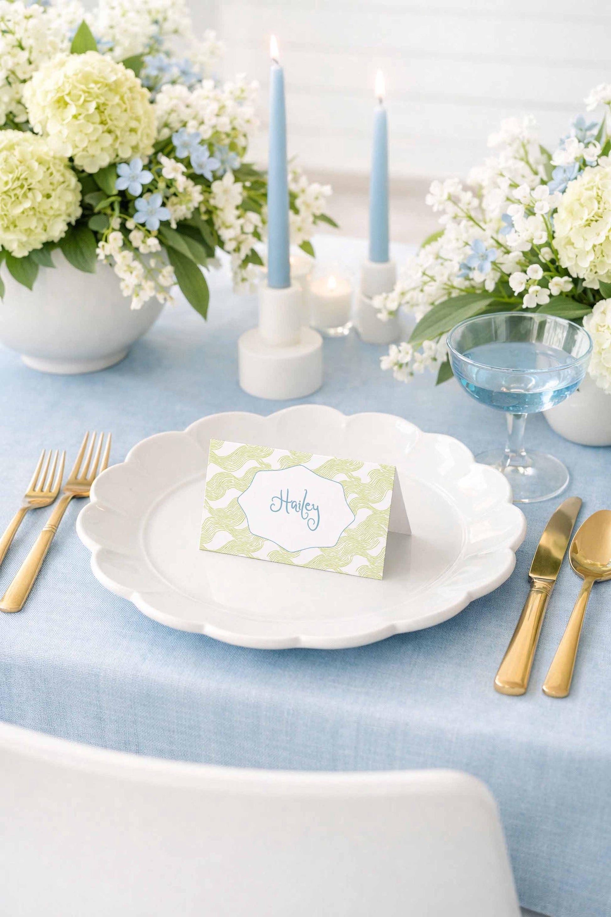 a beautifully set dining table with a white plate, gold cutlery, and a green and white place card with the name "Harley" on it. The table is adorned with white flowers, blue candles, and a glass of water, creating an elegant and inviting atmosphere.