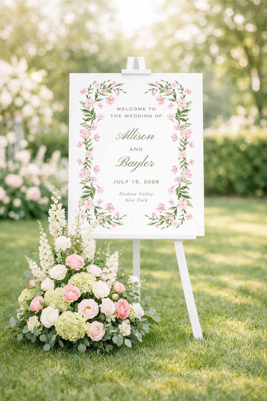 a white wedding sign with pink flowers, standing on a grassy lawn with a floral arrangement in front of it.