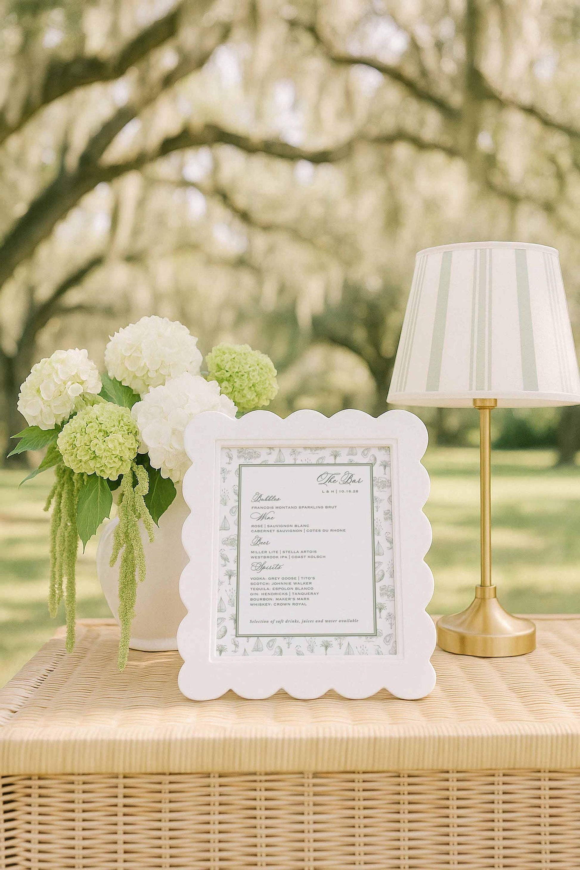 Elegant southern toile bar menu sign shown outdoors on a wicker table, styled with white and green hydrangeas, a striped lamp, and soft daylight filtering through mossy oak trees. The menu features a hand-drawn toile border of palmettos, egrets, oysters, marsh grasses, oak trees, and sailboats. A printable Canva bar sign template perfect for southern weddings, coastal venues, and refined reception drink displays.