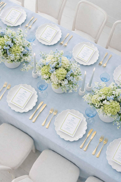 a beautifully set dining table with a light blue tablecloth, white plates, and gold cutlery. The table is adorned with white flowers in blue and white vases, and there are blue and white place cards and glasses arranged on the table.
