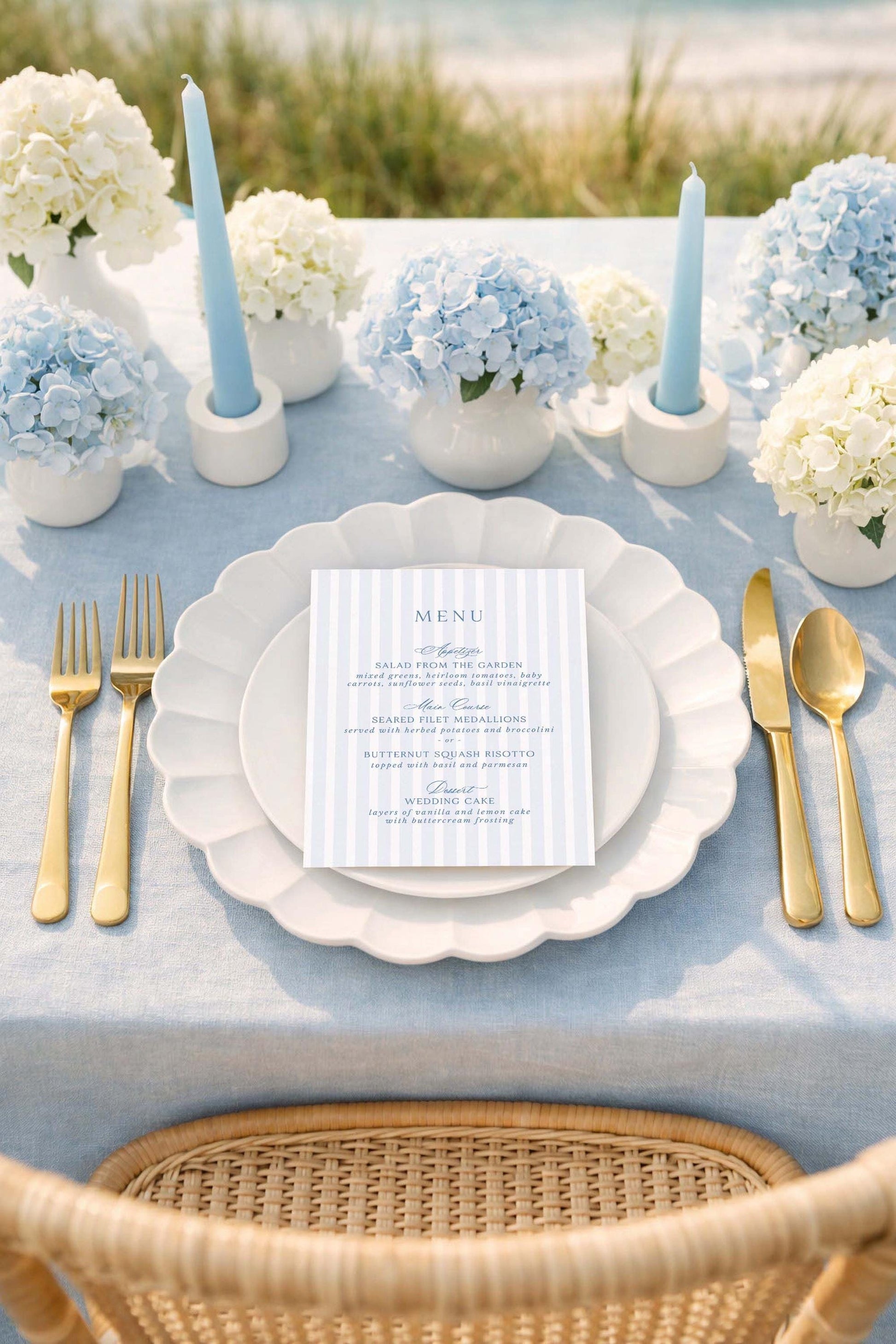 a beautifully set table for a formal dining experience. The table is adorned with a blue and white color scheme, and the centerpiece is a vase filled with blue hydrangeas. The table is set with gold-colored cutlery, including forks, knives, and spoons, arranged neatly around the plates. A menu card is placed on the table, indicating that the table is prepared for a meal. The overall setting suggests a special occasion or celebration, with attention to detail and elegance.