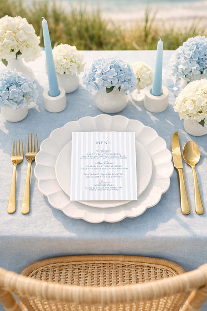 a beautifully set table for a formal dining experience. The table is adorned with a blue and white color scheme, and the centerpiece is a vase filled with blue hydrangeas. The table is set with gold-colored cutlery, including forks, knives, and spoons, arranged neatly around the plates. A menu card is placed on the table, indicating that the table is prepared for a meal. The overall setting suggests a special occasion or celebration, with attention to detail and elegance.