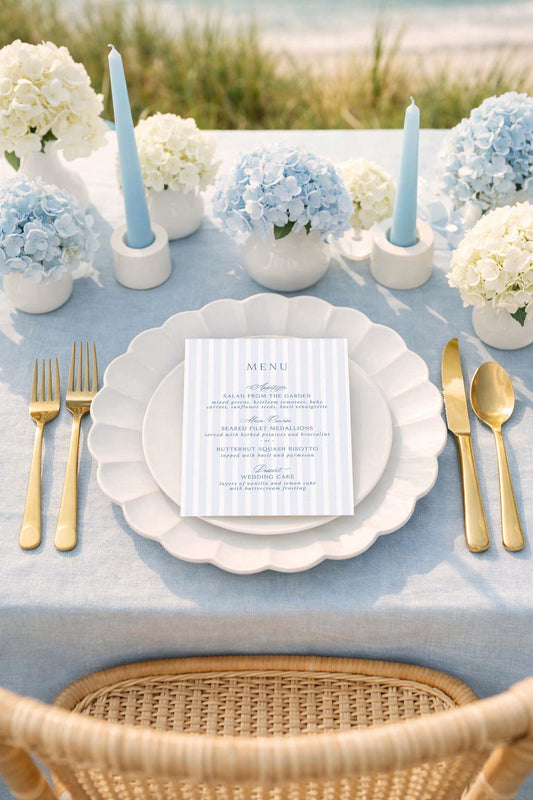 a beautifully set table for a formal dining experience. The table is adorned with a blue and white color scheme, and the centerpiece is a vase filled with blue hydrangeas. The table is set with gold-colored cutlery, including forks, knives, and spoons, arranged neatly around the plates. A menu card is placed on the table, indicating that the table is prepared for a meal. The overall setting suggests a special occasion or celebration, with attention to detail and elegance.