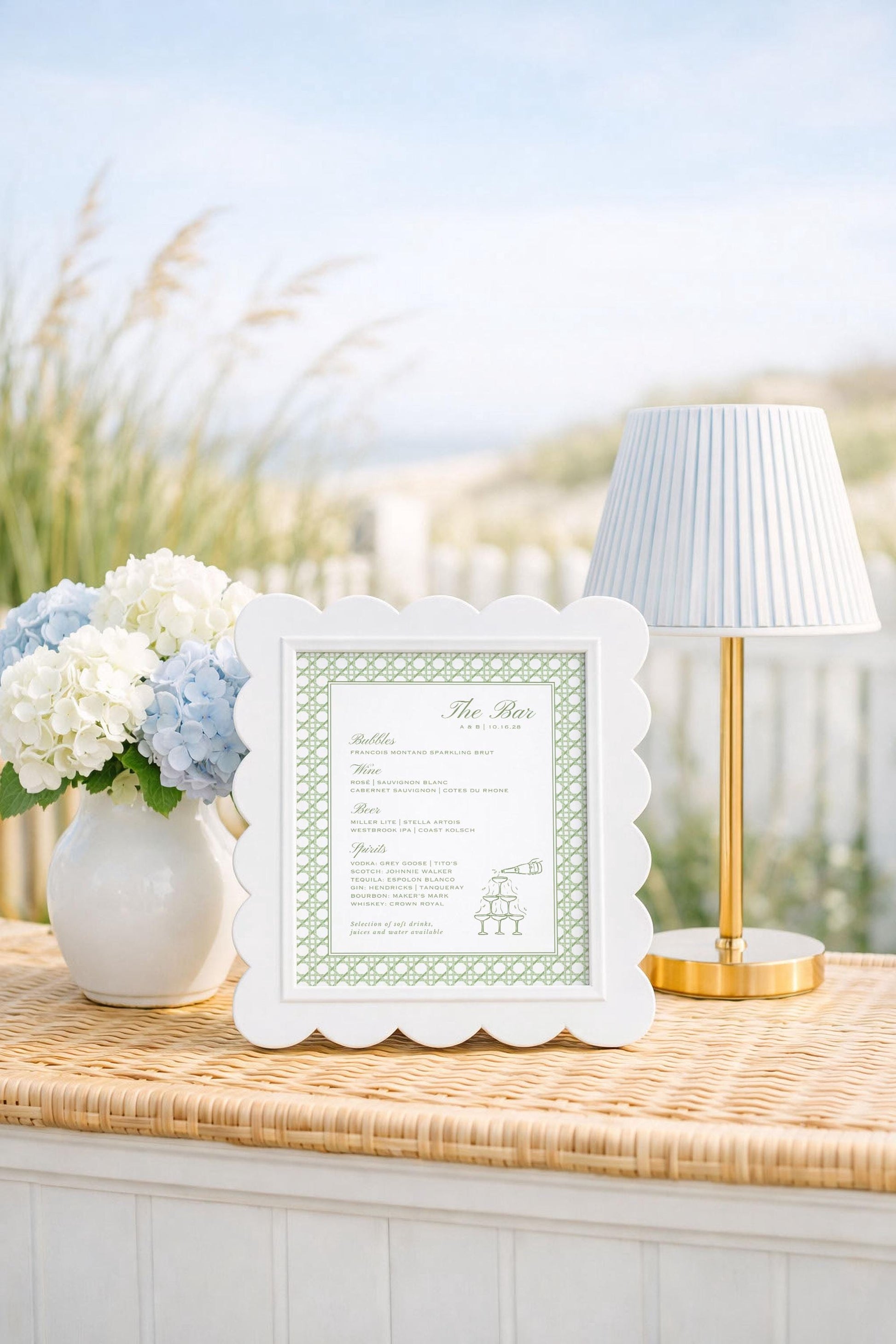 a white frame with a green border, placed on a wicker table, next to a vase of white and blue flowers, and a gold-colored lamp.
