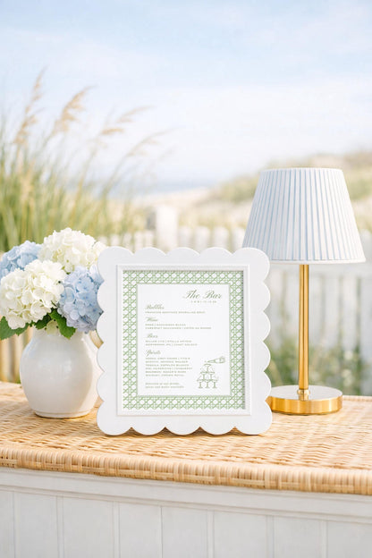 a white frame with a green border, placed on a wicker table, next to a vase of white and blue flowers, and a gold-colored lamp.