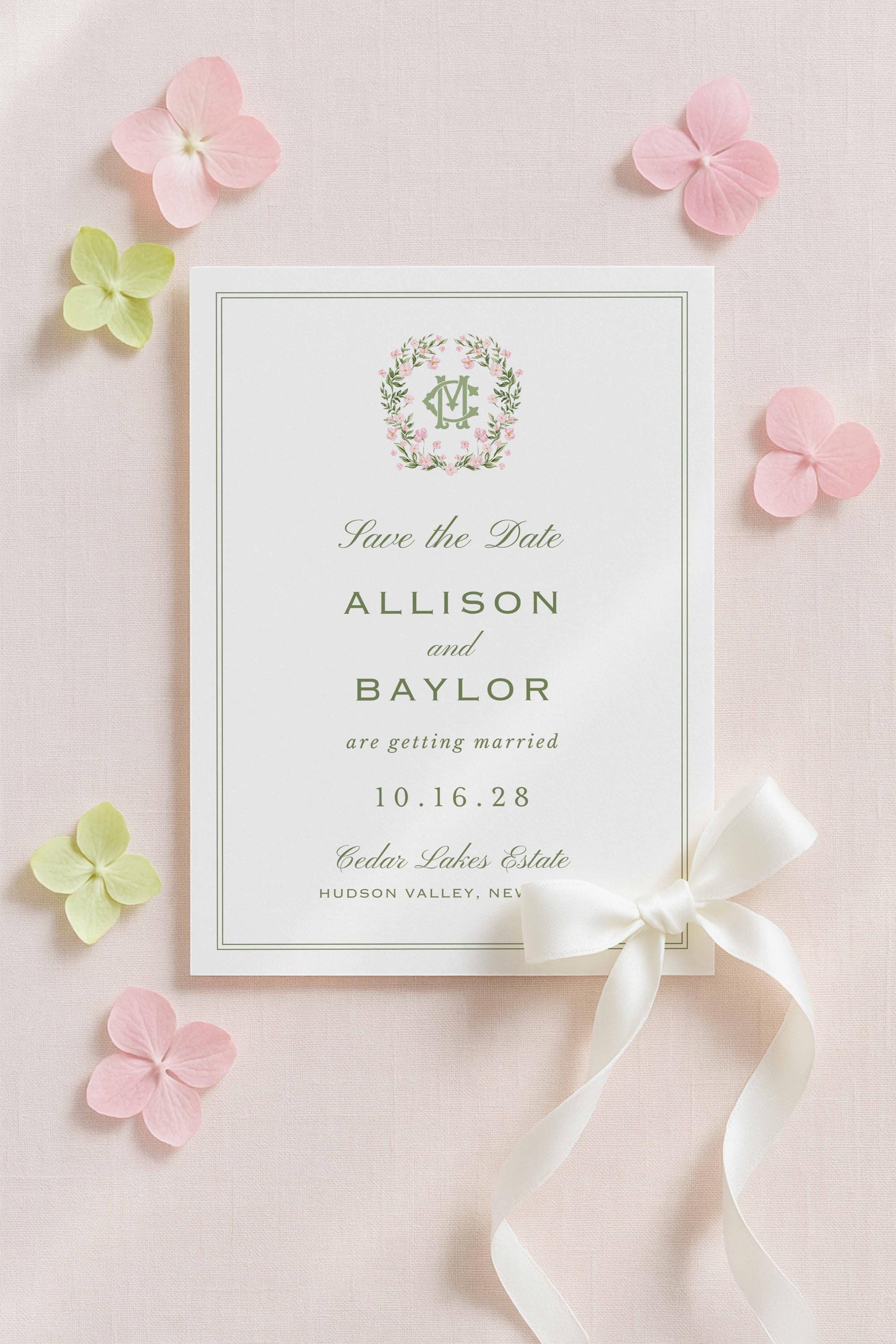 A white wedding invitation card with a green border and floral design, surrounded by pink and green flowers on a light pink background.