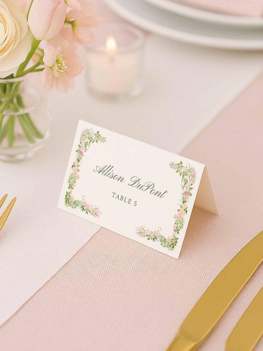 Folded floral escort card displayed at a blush wedding reception table with gold flatware and a pastel floral centerpiece.
