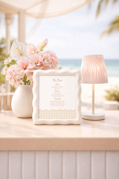 a white table with a framed menu, a vase of pink flowers, and a lamp, all placed on a table with a beach view in the background.