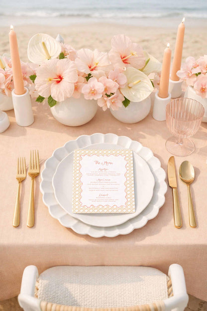 a beautifully set table with a white tablecloth, gold-colored plates, and a menu card. The table is adorned with pink flowers, candles, and glassware, creating an elegant and inviting atmosphere.