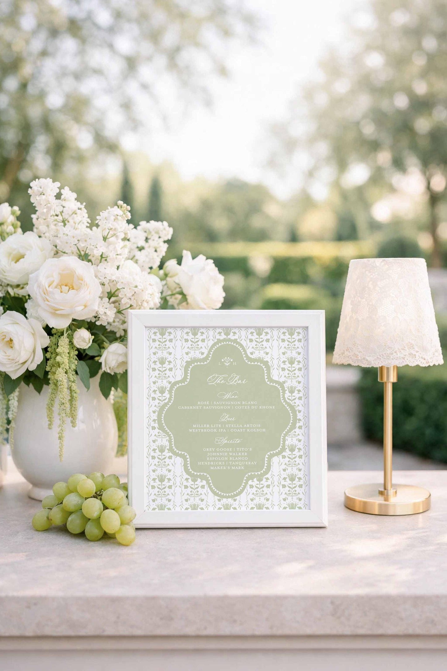 a white framed menu with a floral design, placed on a table next to a vase of white flowers, a bunch of grapes, and a small white lamp.