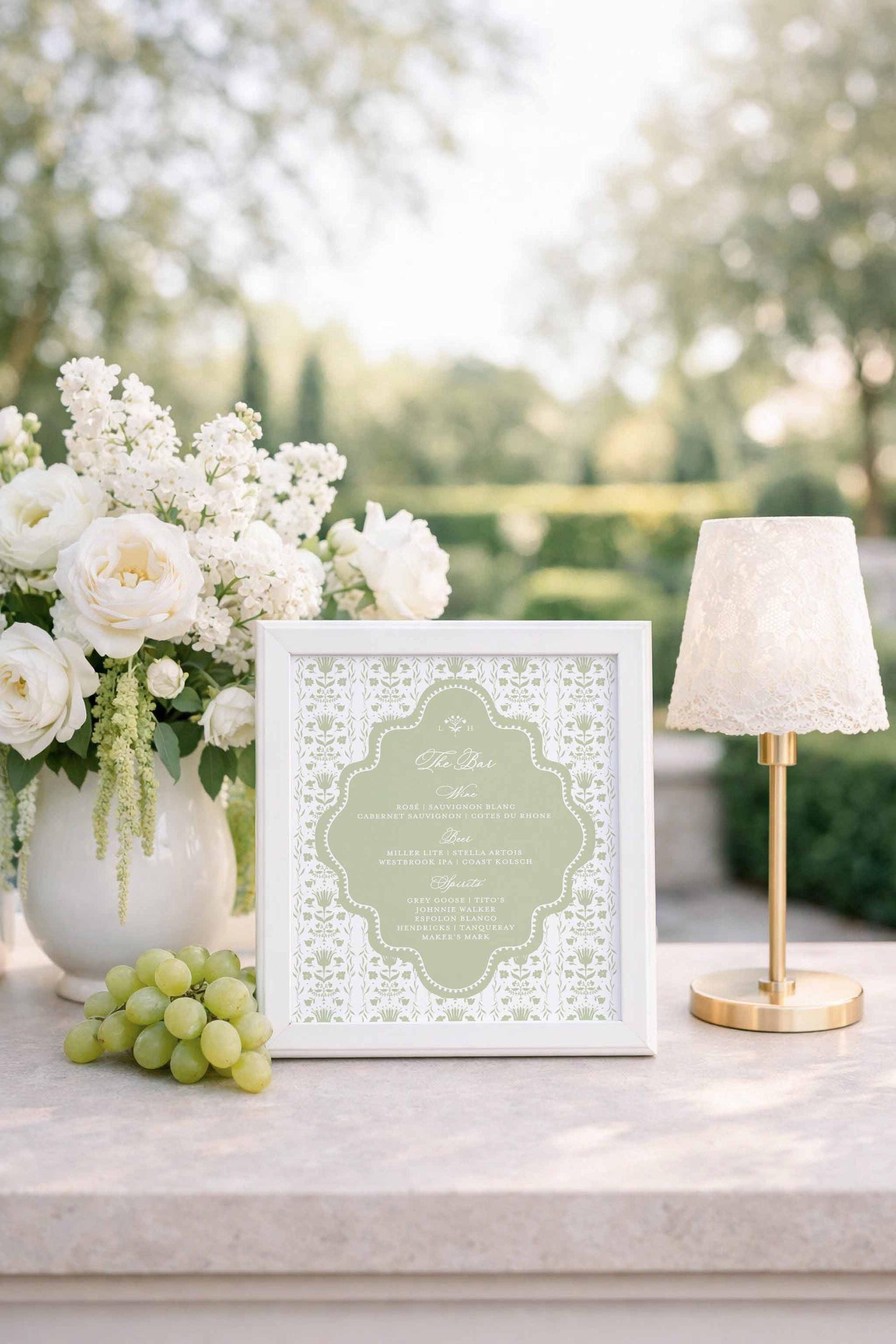 a white framed menu with a floral design, placed on a table next to a vase of white flowers, a bunch of grapes, and a small white lamp.