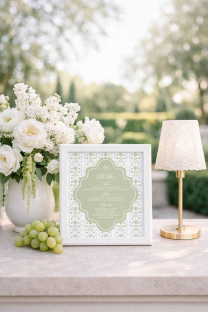 a white framed menu with a floral design, placed on a table next to a vase of white flowers, a bunch of grapes, and a small white lamp.