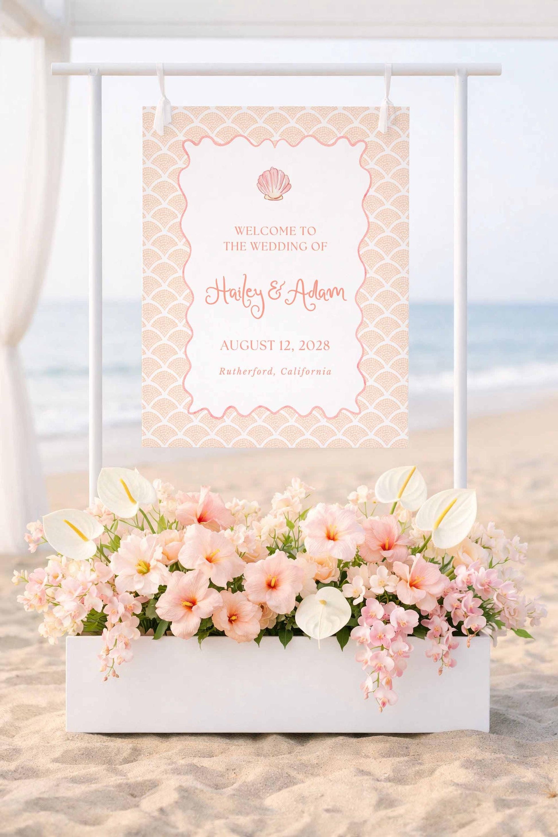 a wedding invitation sign with a beach scene and a white planter filled with pink and white flowers.