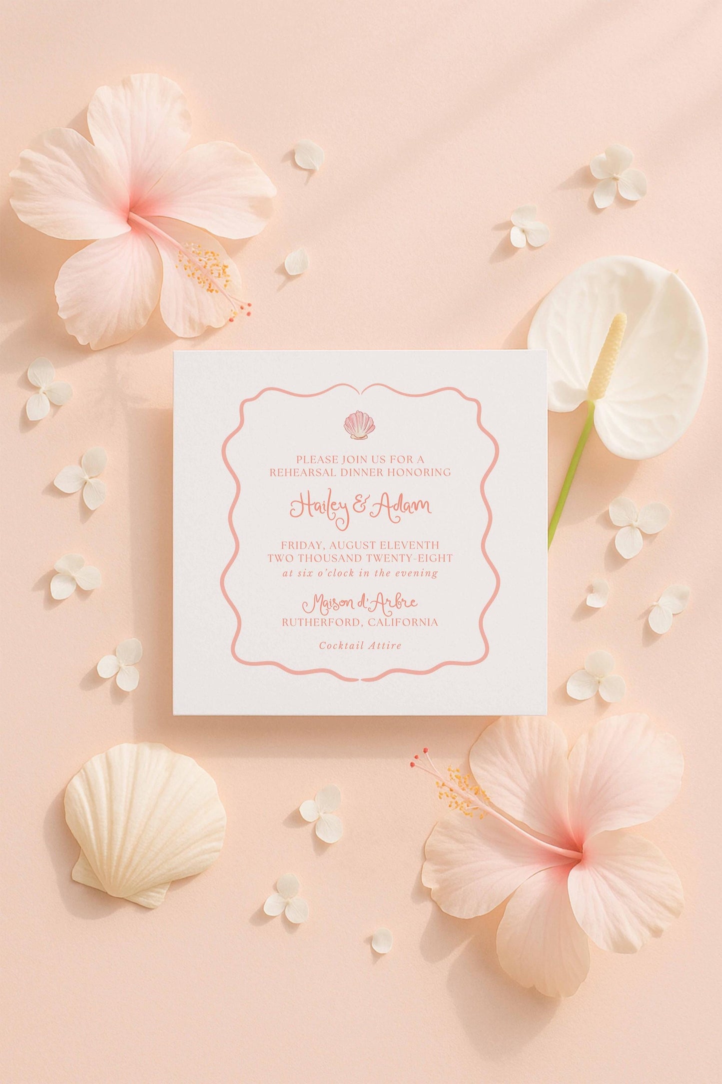 a white wedding invitation card with a pink border, surrounded by various pink and white flowers and shells on a peach-colored background.