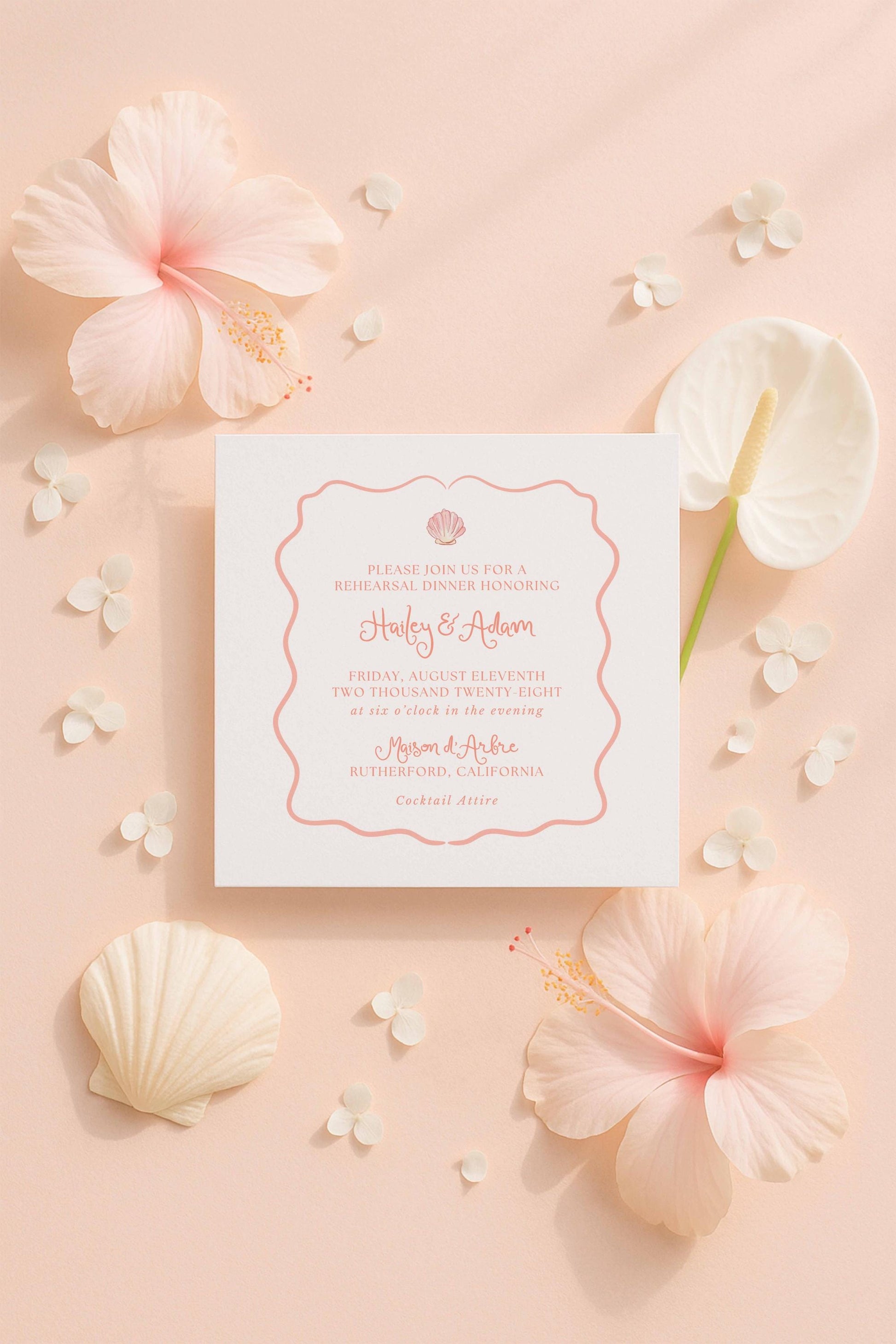 a white wedding invitation card with a pink border, surrounded by various pink and white flowers and shells on a peach-colored background.