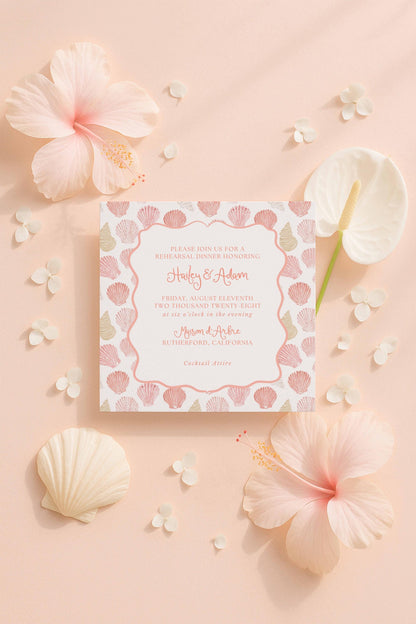 a wedding invitation card with a pink and white floral design, surrounded by various pink and white flowers, shells, and a green pen.