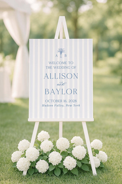 a wedding invitation sign with a white background and blue stripes, placed on a grassy area with white hydrangeas in the foreground.