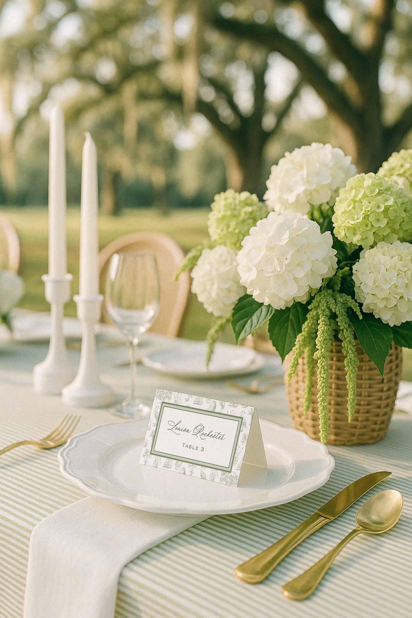 Folded Southern toile wedding escort card displayed on a white scalloped plate at an outdoor reception table. The card features a hand-drawn toile border with palmettos, egrets, oysters, sailboats, marsh grasses, and oak trees. Styled with gold flatware, a white linen napkin, striped green tablecloth, and a woven basket of white and green hydrangeas under oak trees with Spanish moss. Elegant outdoor southern wedding seating card.