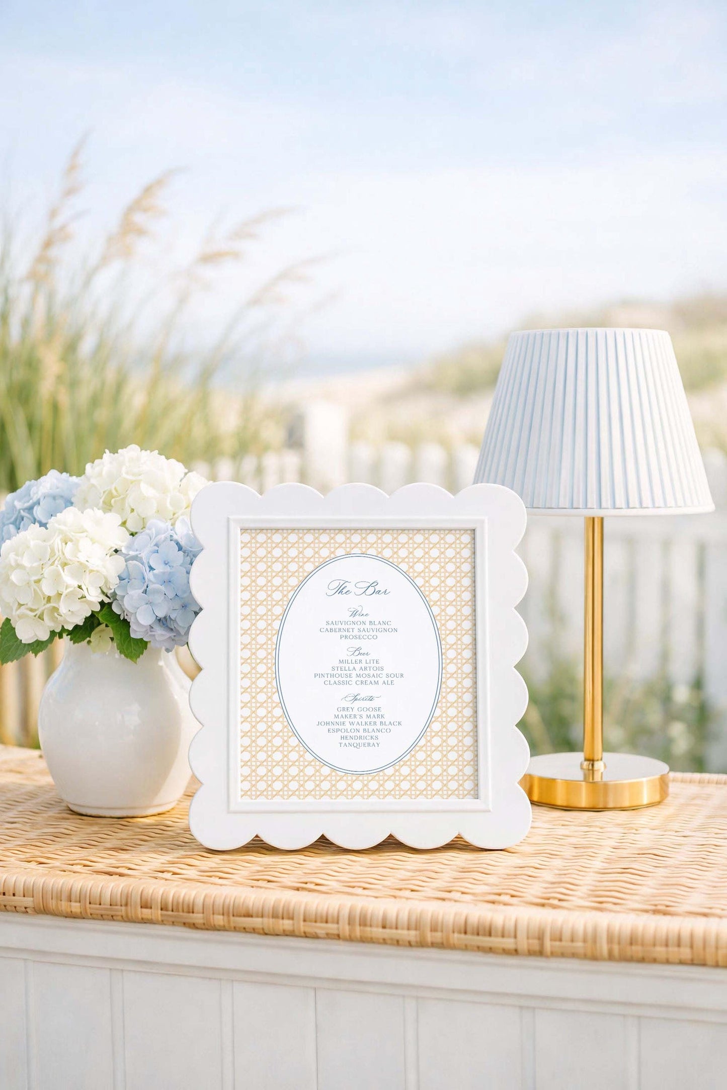 a white frame with a scalloped edge, placed on a woven table, next to a vase of white and blue flowers, and a gold-colored lamp.