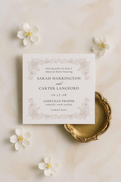 Neutral rehearsal dinner invitation with delicate line-drawn florals layered over a soft beige fabric backdrop and elegant table styling.