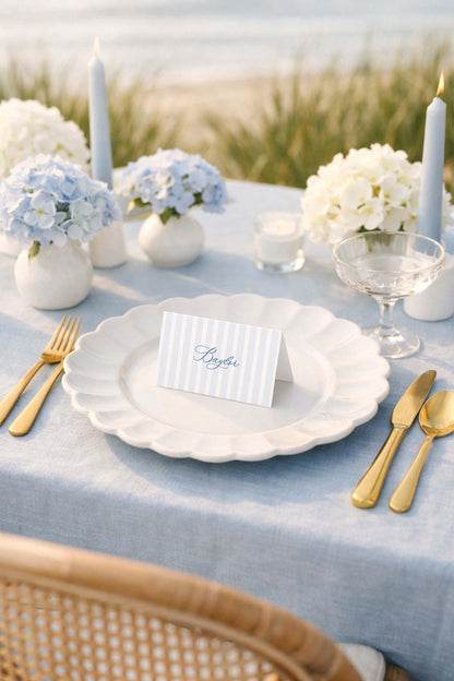 a beautifully set table with a blue tablecloth, featuring a white plate with a name card, gold cutlery, and white flowers.