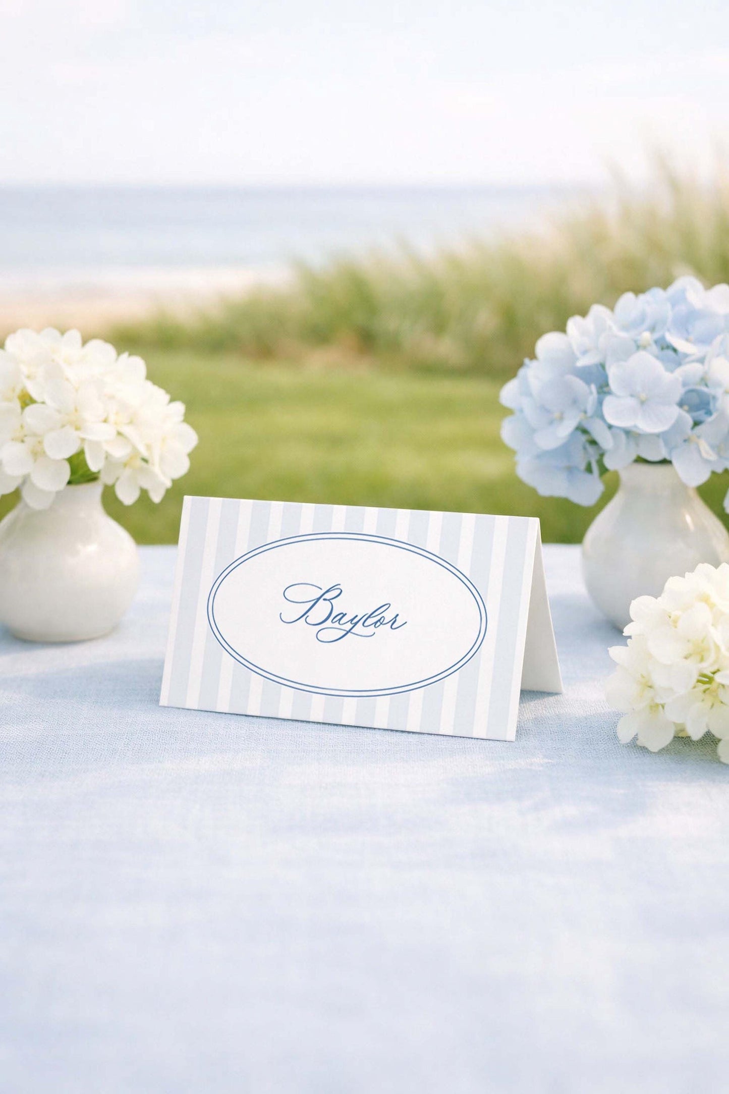 a table with a white tablecloth, a blue and white striped name card, and two vases of white flowers. The name card has the name "Baylor" written on it.