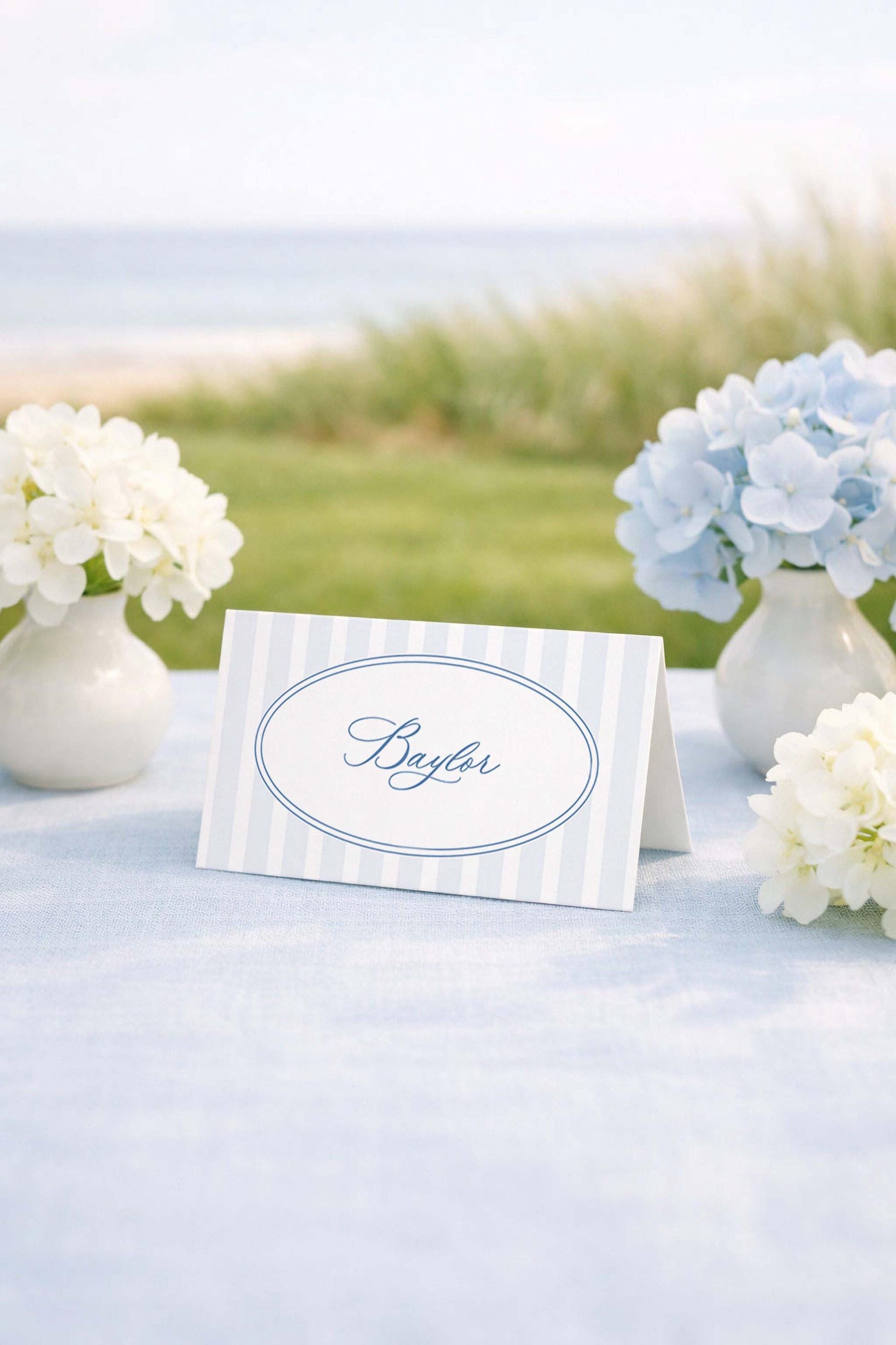 a table with a white tablecloth, a blue and white striped name card, and two vases of white flowers. The name card has the name "Baylor" written on it.