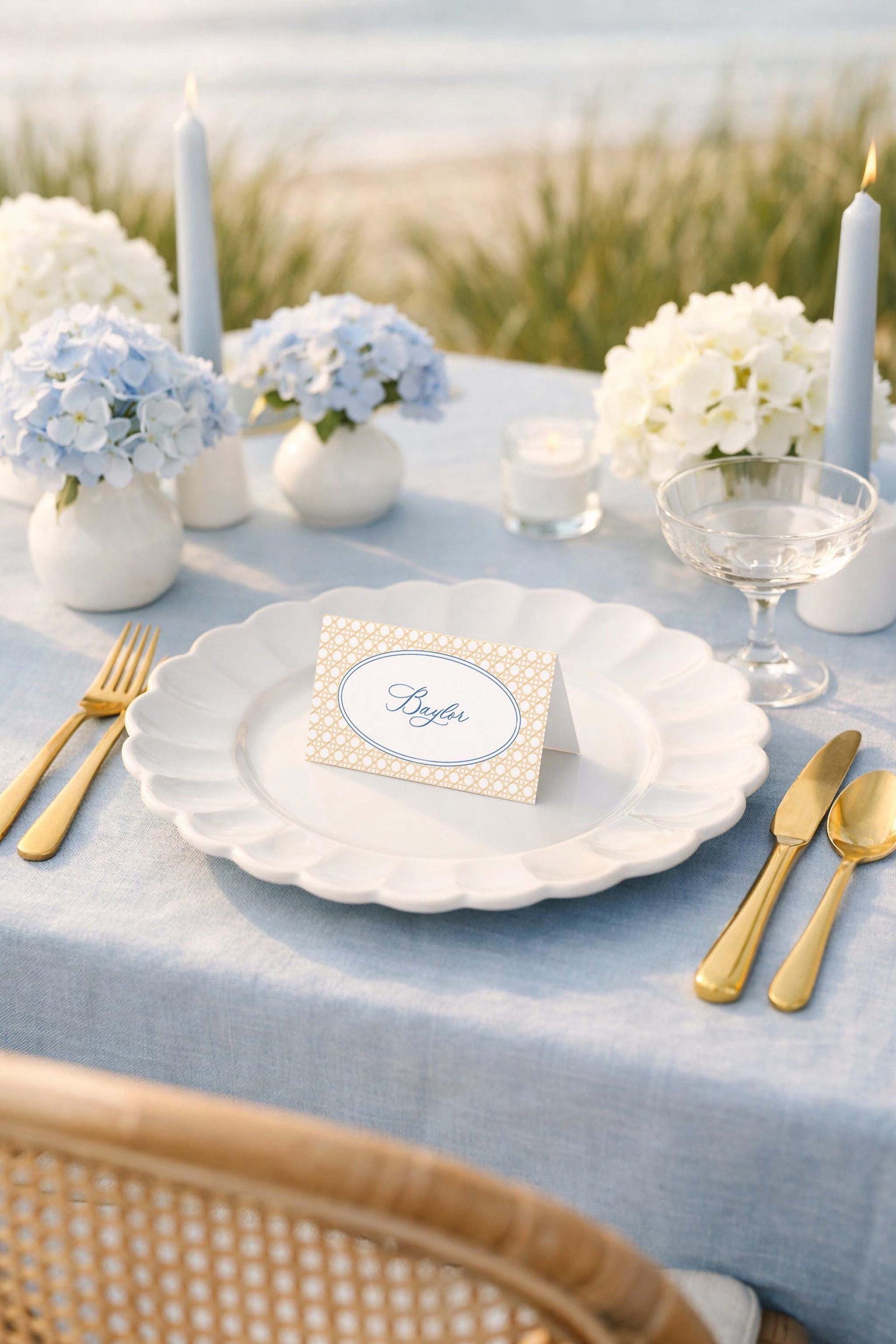 a beautifully set table with a blue tablecloth, white plates, gold cutlery, and a card with the name "Byler" on it. The table is set for a formal dining experience, with a vase of blue hydrangeas and candles as the centerpiece.