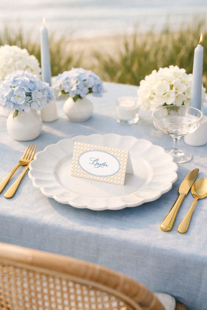 a beautifully set table with a blue tablecloth, white plates, gold cutlery, and a card with the name "Byler" on it. The table is set for a formal dining experience, with a vase of blue hydrangeas and candles as the centerpiece.