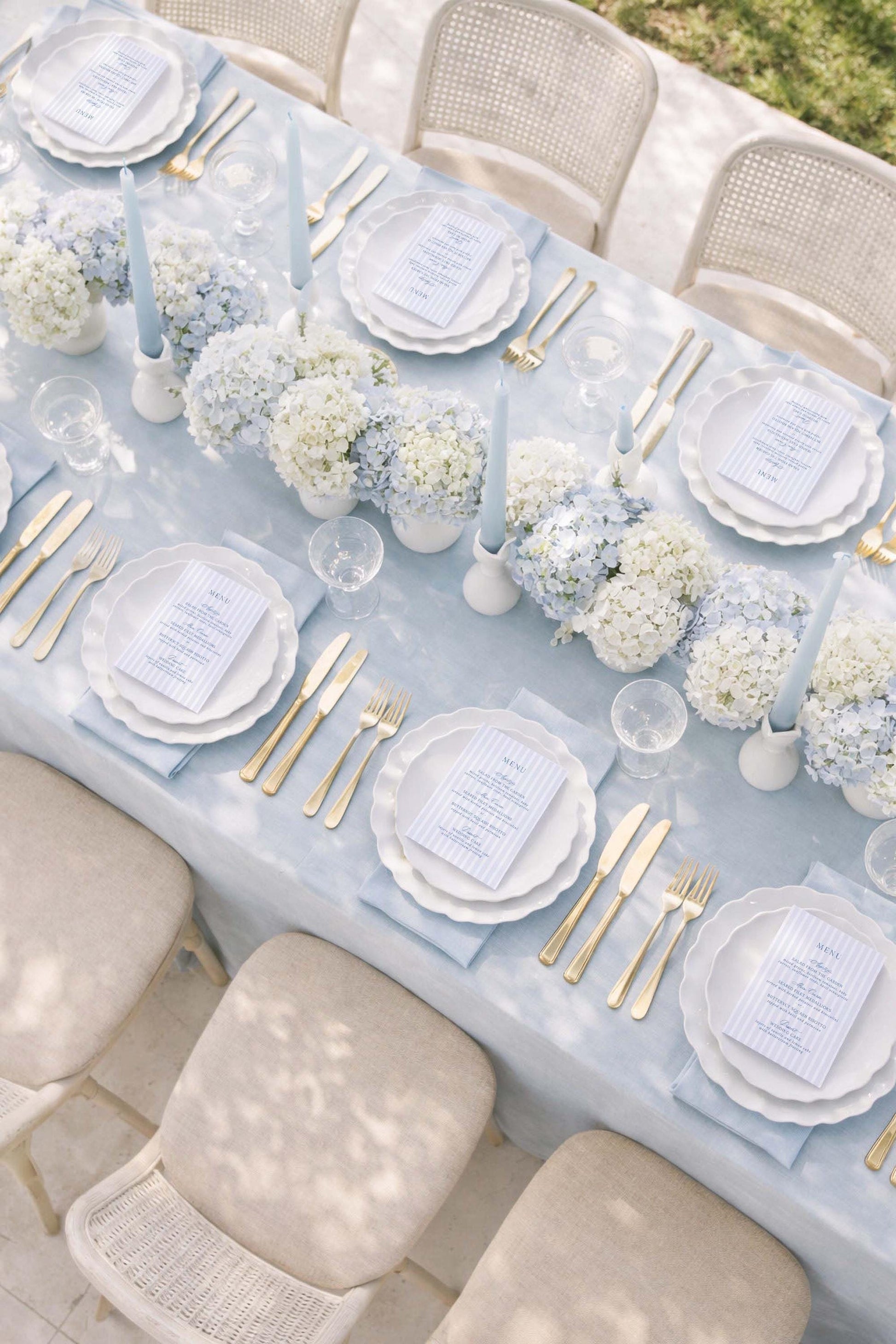a beautifully set dining table with white plates, gold cutlery, and a floral centerpiece. The table is surrounded by beige chairs, and the table is set with candles and place settings.