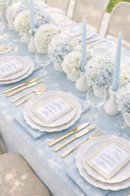 a beautifully set dining table with white plates, gold cutlery, and elegant floral arrangements. The table is set for a formal event or celebration, with a menu card placed in front of each plate.