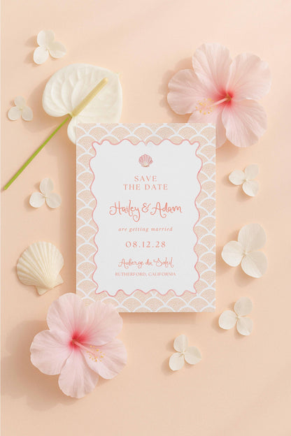 a white wedding invitation card with a pink and white floral design, surrounded by various pink and white flowers and shells on a peach background.