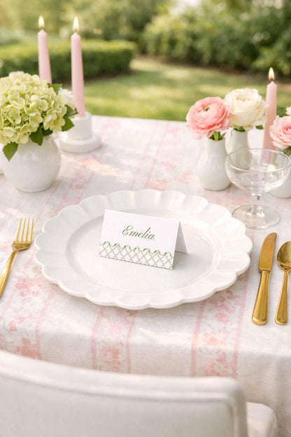 a beautifully set table with a white tablecloth, pink flowers, and a white plate with a name card. The table is set for a formal dinner or event, with gold cutlery and a vase of flowers.
