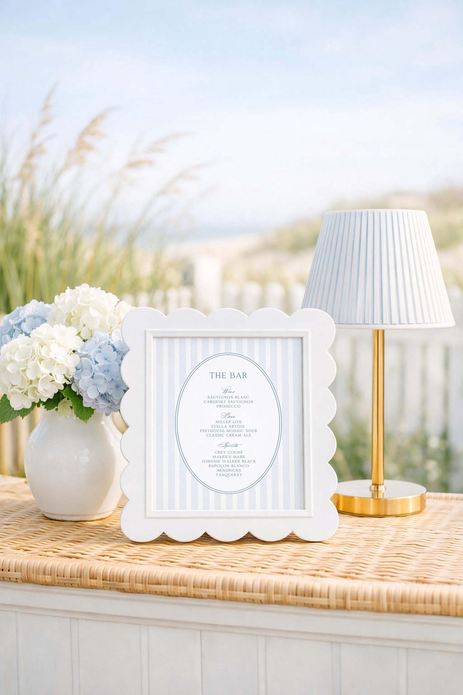 a white bar sign with blue stripes, a white vase with white and blue flowers, and a white lamp with a gold base, all placed on a woven table.