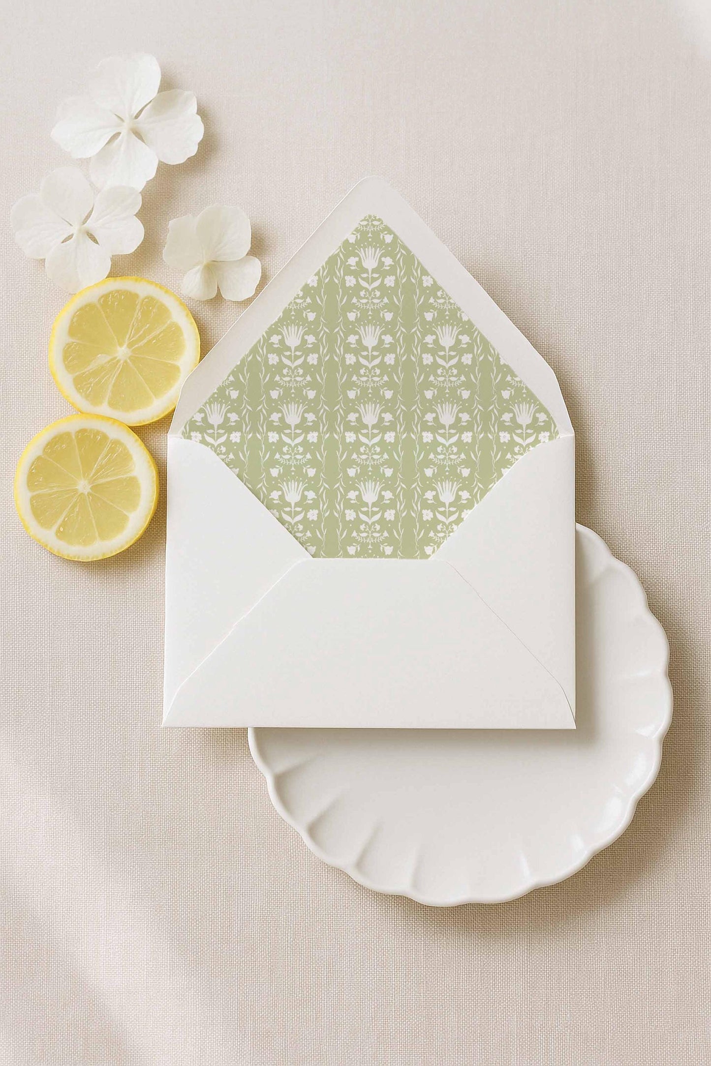 a white envelope with a green floral pattern, placed on a white plate, along with two lemon slices and white flowers.