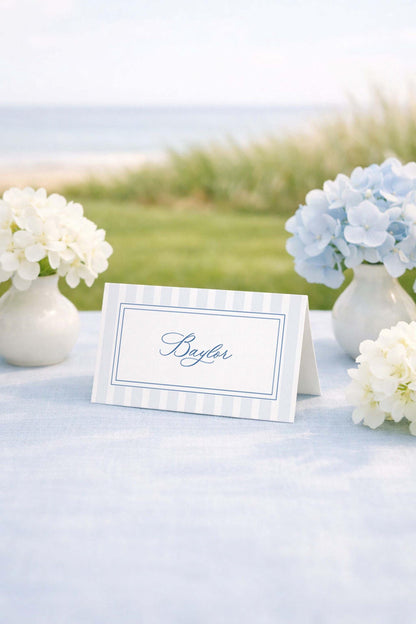 a table with a white tablecloth, a white card with the name "Baylar" written on it, and two vases of white flowers. The table is set against a backdrop of a grassy field and a body of water, creating a serene and picturesque setting.