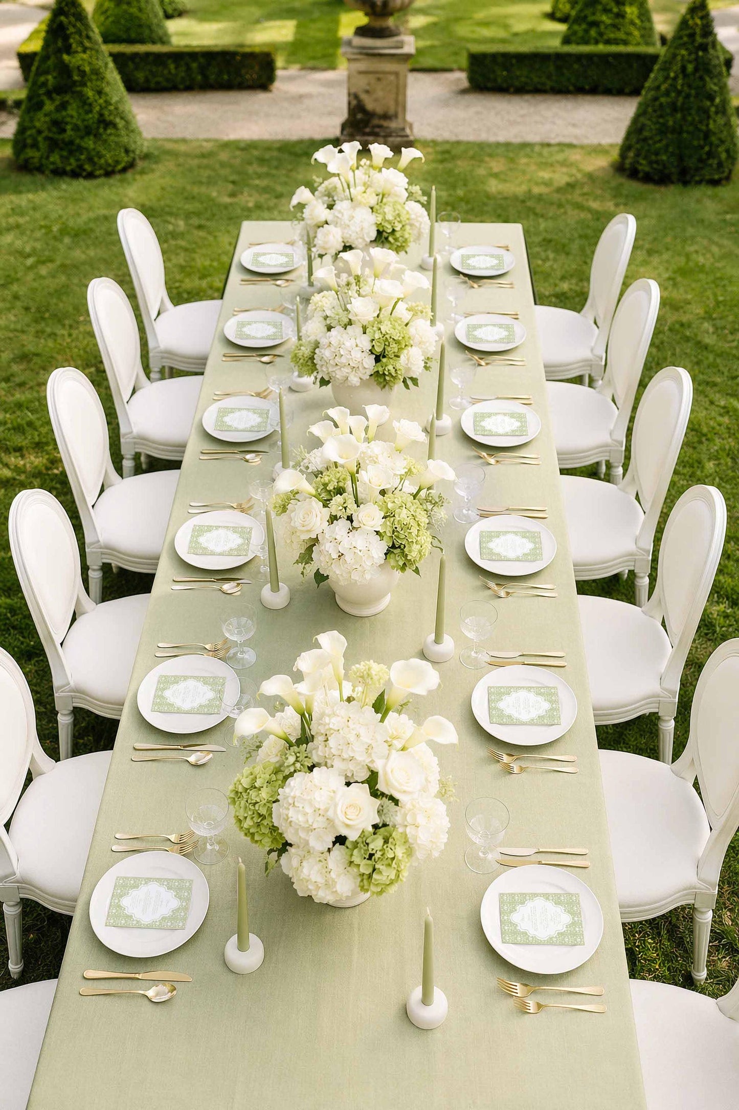 a beautifully set outdoor dining table with white chairs, white plates, and white flowers arranged in vases. The table is set for a formal event or celebration, with elegant tableware and floral centerpieces.