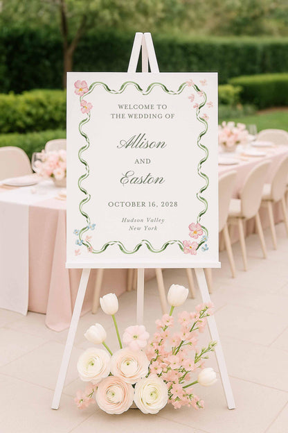 Printable hand-painted watercolor wedding welcome sign with lush pink and green floral frame on a white easel surrounded by elegant blush flowers and outdoor reception decor.