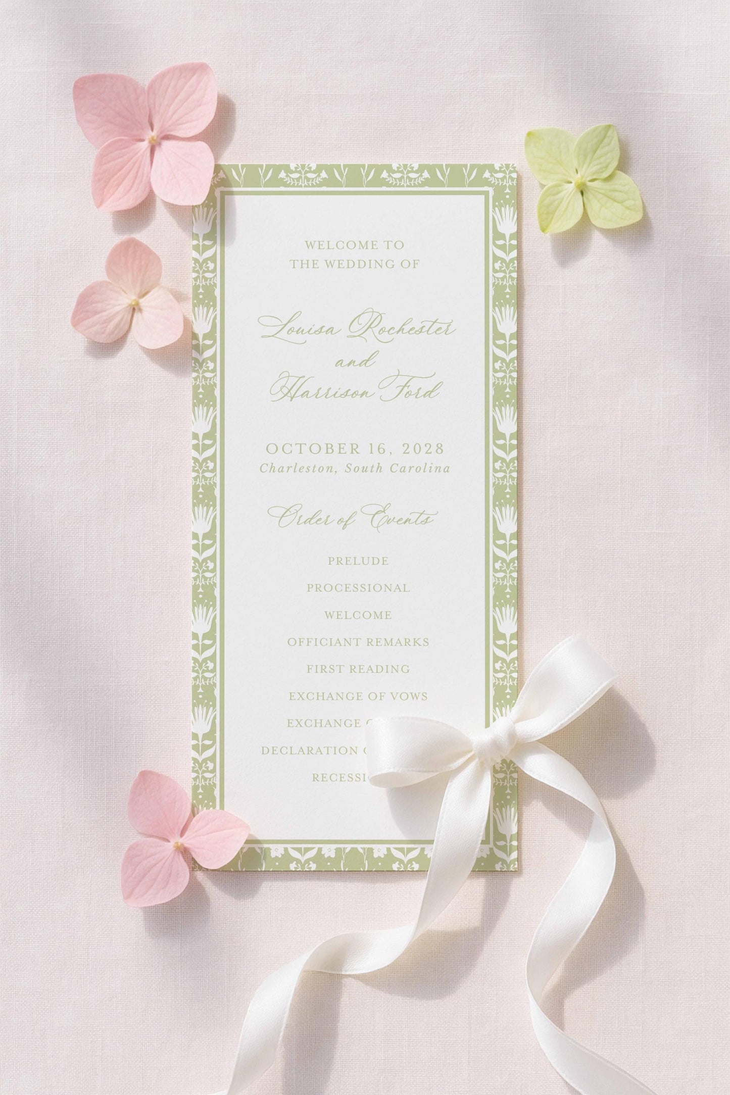 a wedding invitation card with a green and white color scheme, adorned with pink and green flowers. The card is placed on a light pink background, and there are additional flowers scattered around it.