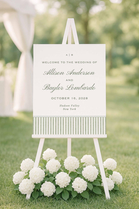 Green striped watercolor welcome sign displayed on an easel surrounded by white flowers outdoors.